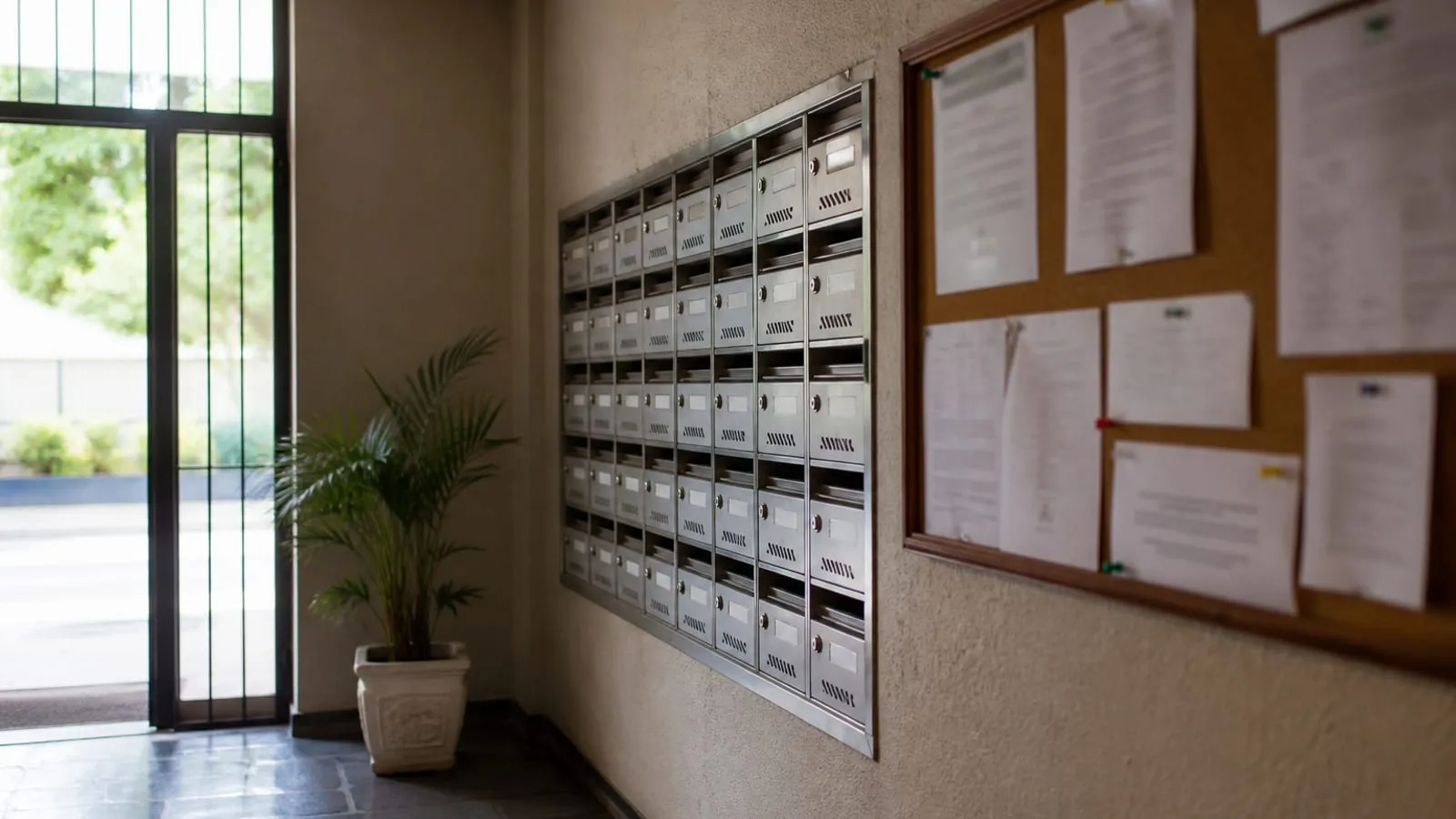 Hall de condomínio com mural de avisos, caixas de correio metálicas e uma planta junto à entrada